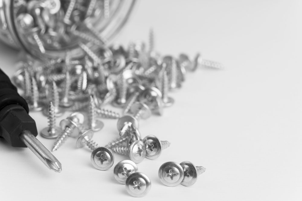 Dezenas de parafusos de cabeça chata arredondada e de aço inox caídos de um pote de vidro ao lado de uma chave de fenda preta.