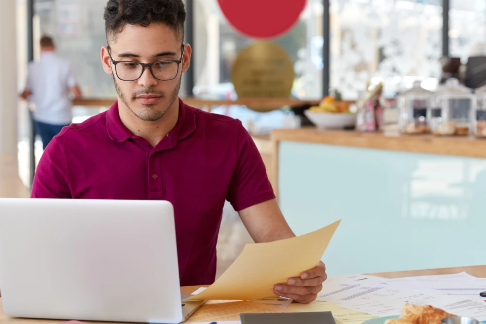 Um homem assistente de compras júnior com vários papéis na mão em frente a um notebook tentando saber como entender um orçamento de fixadores.